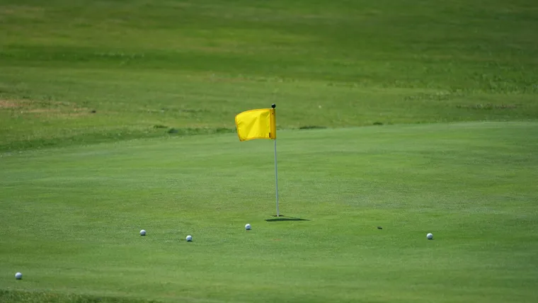 A closely cut putting green in bright sun with a yellow flag in the cup and several golf balls scattered nearby.