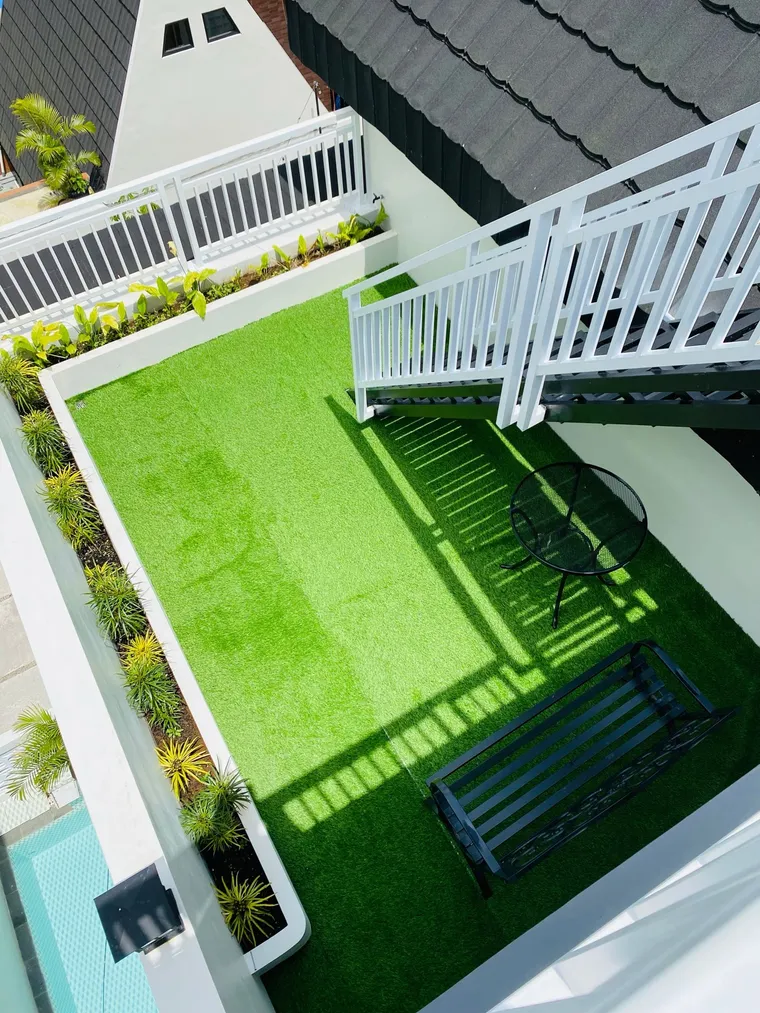 A small rooftop terrace laid with turf, enclosed by white railings, with tropical planting, a bistro table and a bench.